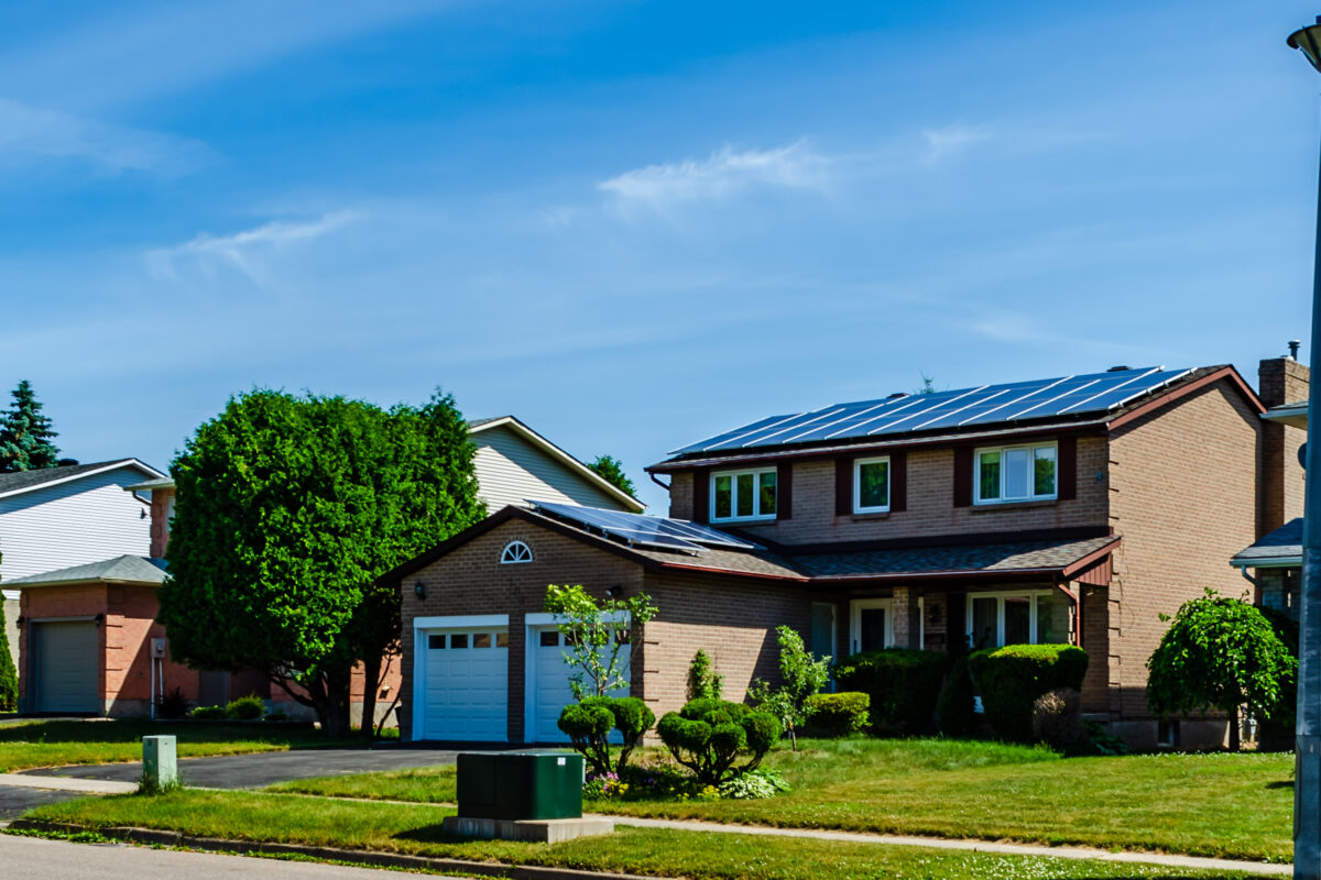 Panorama of a row of residential houses along a street, one with solar panels on the roof under a blue sky